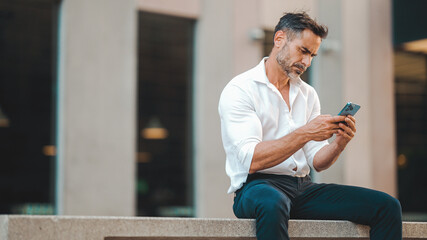 Mature businessman with neat beard uses mobile phone sits on bench in the financial district in the city. Successful man scrolls through information on smartphone and smiles