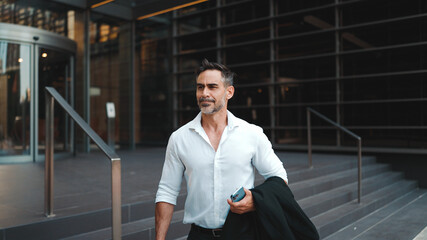 Mature businessman with neat beard wearing white shirt leaves the office in the financial district in the city. Successful man with mobile phone in his hands, after hard day's work