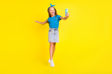 Full size photo of excited small girl jumping hold telephone make selfie photo video call isolated on yellow color background