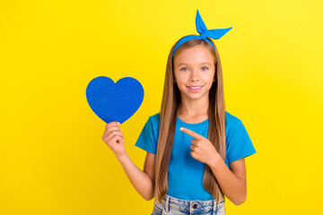 Photo of small cheerful schoolgirl direct finger hand hold paper heart card isolated on yellow color background