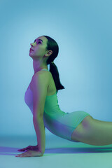 Portrait of young sportive girl doing stretching exercises isolated over blue studio background in neon light. STrong back exercises