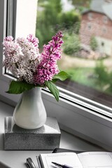 Cozy home still life: spring lilac flowers in a white vase on the windowsill, notebook and glasses. Retro style, vertical photography