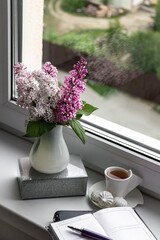 Still life with lilac flowers in a white vase on the windowsill. Notepad for notes and a cup of coffee, cozy home business planning