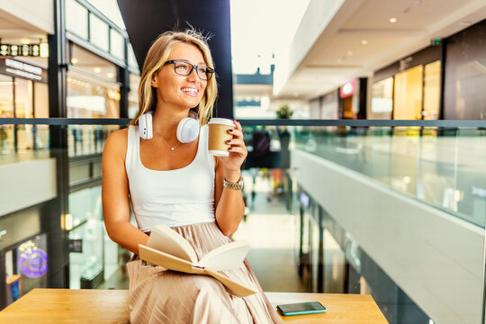 Shot of a senior woman enjoying the coffee while reading a book, outdoor. Young beautiful woman in glasses reading interesting book in the shopping mall.