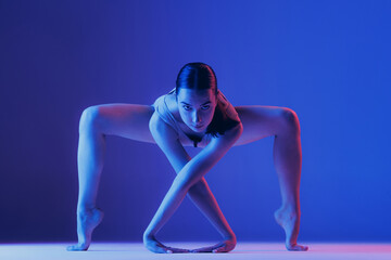 Portrait of young sportive girl doing stretching exercises isolated over blue studio background in neon light. Standing on tiptoe