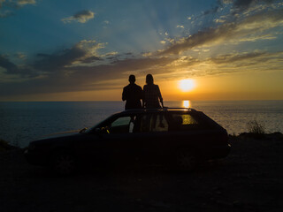 Silhouette of a happy couple sitting on the roof of a car on a cliff in front of the sea with a beautiful orange-blue sky at sunset, view from a drone. Concept of vacations and travel, romance - Powered by Adobe