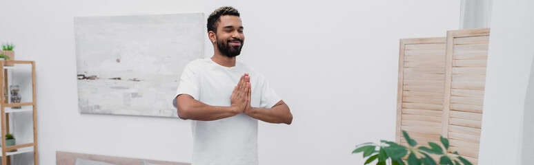 Fototapeta na wymiar happy african american man african american man meditating with praying hands near folding screen, banner.