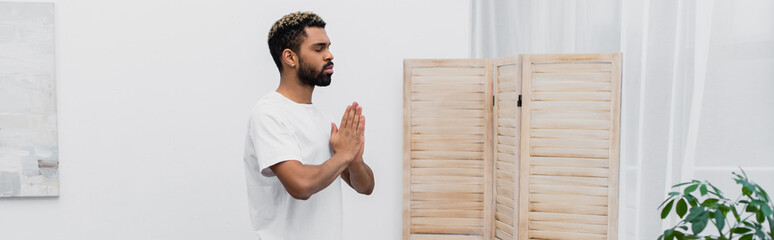 bearded african american man meditating with praying hands near folding screen, banner.