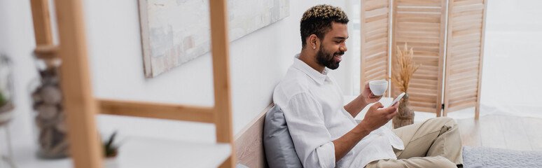Fototapeta premium happy african american man with dyed hair holding cup of coffee and using smartphone while resting on bed, banner.