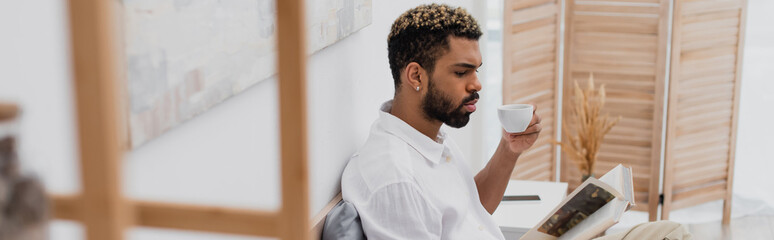 Fototapeta premium young african american man with dyed hair reading book and holding cup of coffee in modern bedroom, banner.