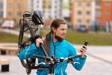 Fototapeta premium A confident young bearded man is carrying his bicycle on his shoulder and looking at a smartphone while walking outdoors. Stylish businessman with a bike.