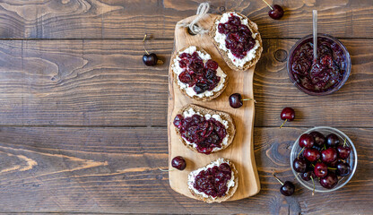 Wholewheat toasts with ricotta and homemade sweet cherry jam on wooden board and background. Copy...