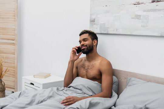 Shirtless And Happy African American Man With Dyed Hair Talking On Mobile Phone In Bed.