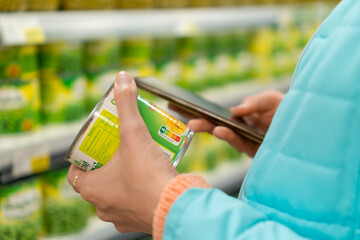 Close-up of the hands of a young woman holding a can of canned corn in a store with a mobile phone in her hands. Concept of buying food, proper nutrition - obrazy, fototapety, plakaty