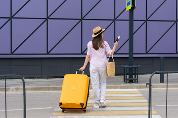 Fototapeta na wymiar Woman in a straw hat shirt and pants with a passport and tickets in her hands and a straw bag an orange suitcase crosses a pedestrian crossing goes near the airport. Concept: travel 