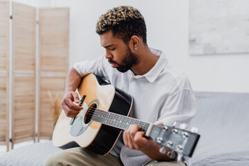 focused young african american man with dyed hair playing acoustic guitar in bedroom.