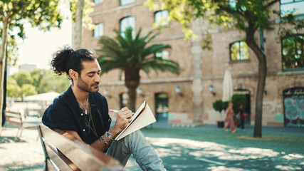 Young italian guy with ponytail and stubble sits on street bench and makes sketches with pen on piece of paper