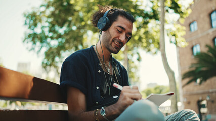Young italian guy with ponytail and stubble sits with headphones on street bench listens to music...