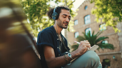 Young italian guy with ponytail and stubble sits with headphones on street bench listens to music and makes sketches with pen on piece of paper on cityscape background - obrazy, fototapety, plakaty