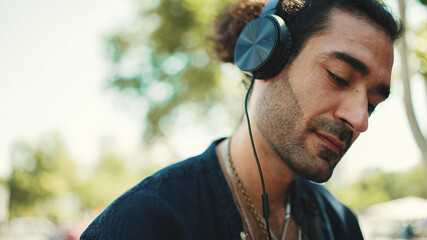 Closeup of young italian guy with ponytail and stubble sits with headphones on street bench and...