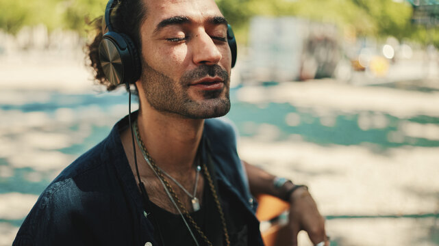 Fototapeta Closeup of young italian guy with ponytail and stubble sits with headphones on street bench and listens to music on cityscape background