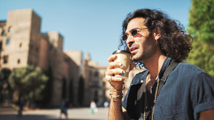 Young italian guy with long curly hair and stubble is enjoying aromatic coffee. Stylish man drinks...