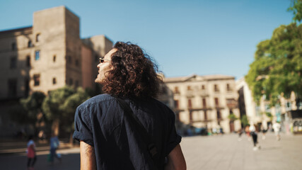 Young italian guy with long curly hair and stubble walks along the fenced sidewalk and looks around.