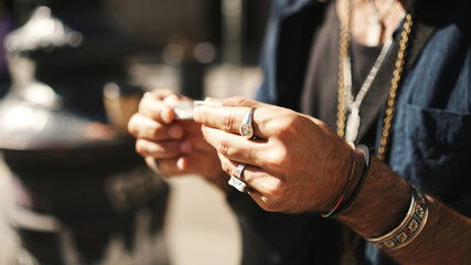 Close-up of male hands in bracelets and rings rolls cigarette. Male hands wrapping tobacco in...