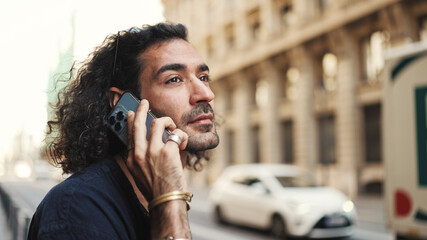 Young attractive italian guy with long curly hair and stubble is using mobile phone. Stylish man with talking on smartphone on busy street