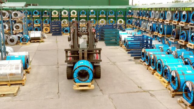 The forklift moves through the warehouse under the open sky. Transports rolls of metal tiles. Products are neatly stacked around on the shelves.