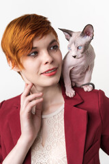 Portrait of hipster redhead young woman with short hair, dressed in red jacket and playful Sphynx Cat sitting on her shoulder. Studio shot on white background. Part of series.