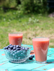 Two glasses with strawberry smoothie, bowl of blueberry on green wooden table. Healthy breakfast in the garden. Eating fresh concept. 
