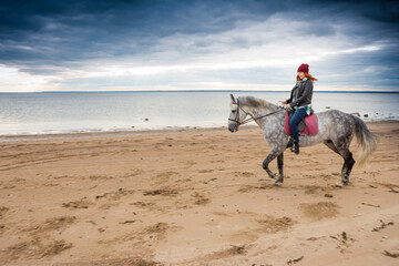 dressing jeans, jacket and spring hat woman rides on horseback along sandy beach