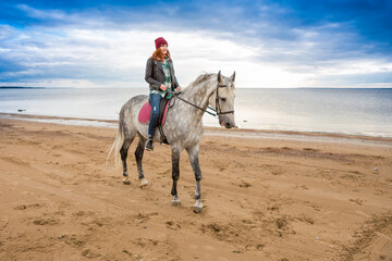 dressing jeans, jacket and spring hat female horseback raider gallops on a dappled hack along sea-coast