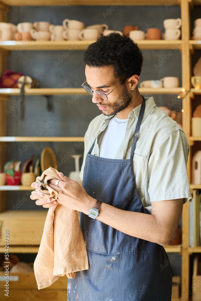 Poster young multi-ethnic craftsman in apron wiping his hands with towel while standing against display wit