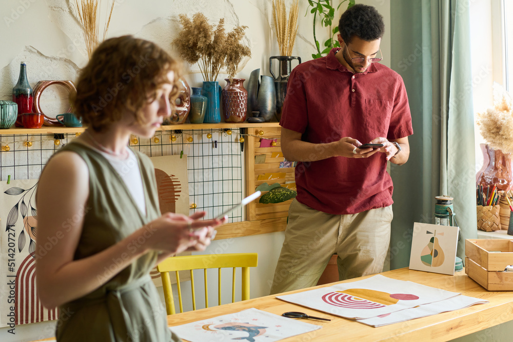 Poster Young multi-ethnic man with smartphone taking photo of drawing on paper while standing by table with his colleague using tablet in front