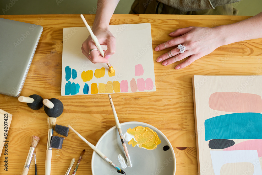 Poster above view of hands of young creative woman making color swatches on paper while sitting by table an