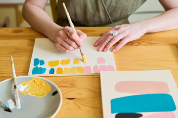 Obraz premium Hands of young woman with paintbrush making color swatches for her new painting while sitting by wooden table in workshop