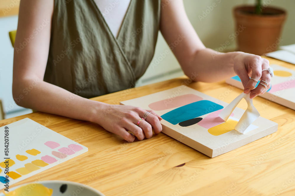 Poster hands of young creative female sitting by wooden table in workshop or studio and using stencil while