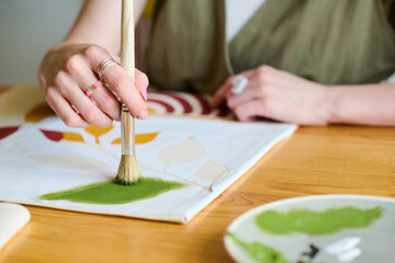 Hand of young creative female artist with paintbrush putting green watercolor on paper while...