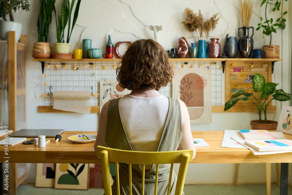 Aufkleber Rear view of young creative woman sitting by table in workshop with handmade souvenirs on shelf along wall and creating new paintings