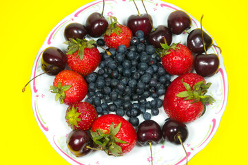 Strawberries, Sweet wild cherries and Blueberries on a plate on the yellow background. Delicious and fresh summer fruits. Healthy vegetarian food with vitamins.