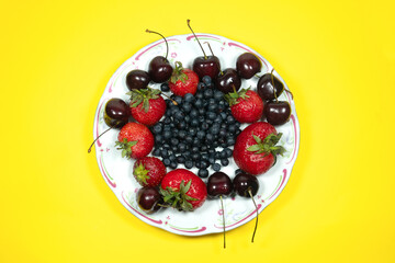Strawberries, Sweet wild cherries and Blueberries on a plate on the yellow background. Delicious and fresh summer fruits. Healthy vegetarian food with vitamins.