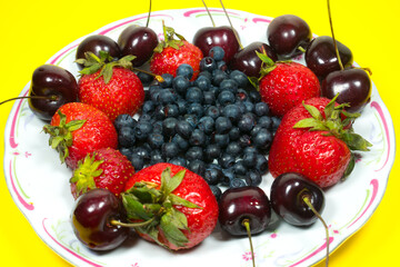Strawberries, Sweet wild cherries and Blueberries on a plate on the yellow background. Delicious and fresh summer fruits. Healthy vegetarian food with vitamins.