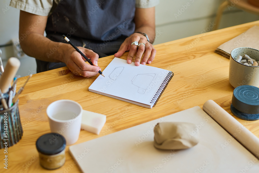 Poster hands of young creative craftsman making sketch of cup on page of notepad while sitting by table wit