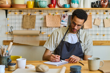 Young creative black man drawing sketches of new handmade craft items in notepad or making working notes while sitting by table in studio