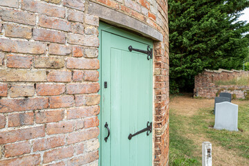 Exterior view of a very old round house, once a dove house. Showing the newly painted wood door. Seen in a rural graveyard.