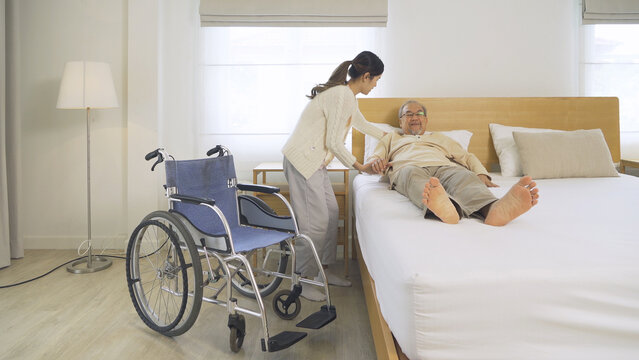 Asian woman nurse, daughter help father to get up from bed to wheelchair, supporting old senior elderly patient in bedroom in home or house in healthcare. People lifestyle. Family disability therapy.