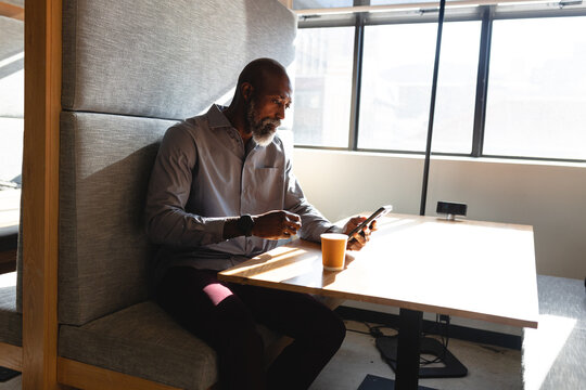 African american mature businessman using smart phone while having coffee during break in office