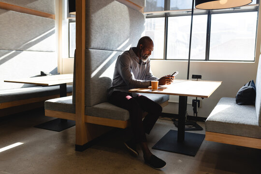 African american mature businessman using mobile phone while having coffee during break in office